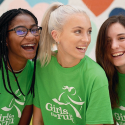 A candid photograph of three girls standing side-by-side, smiling and laughing joyfully. They are wearing bright green Girls on the Run t-shirts.
